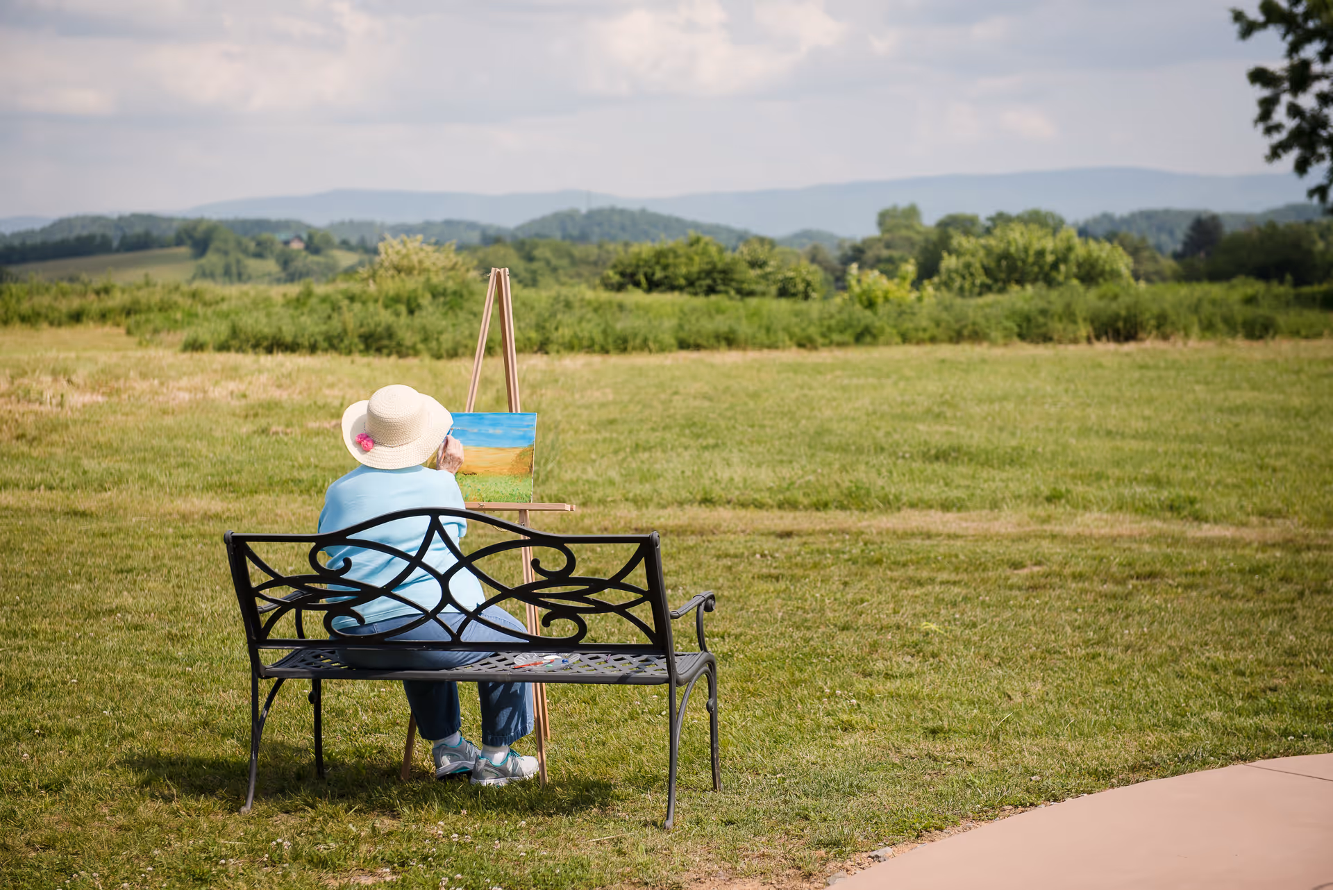 Senior woman painting outdoors on a grassy field beside a decorative bench.