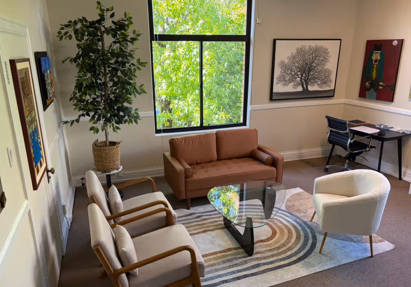 A cozy senior living lounge with sofa, armchairs, glass coffee table, large window, and plants.