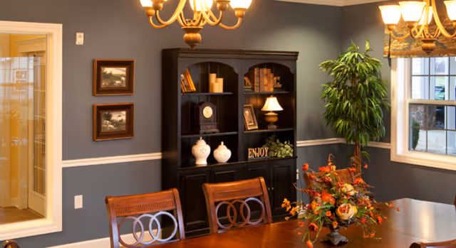 Dining room with a wooden table and chairs, a black hutch with decorative items, chandelier, and a windowed wall with a potted plant.