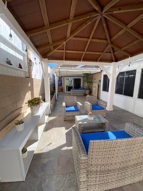 Covered outdoor patio area with a wooden roof structure, featuring wicker chairs with blue cushions arranged around a glass-top table. There is a white shelving unit with potted plants on the left side and string lights hanging from the ceiling. The space is enclosed by walls with windows on the right side and a sliding glass door at the far end.