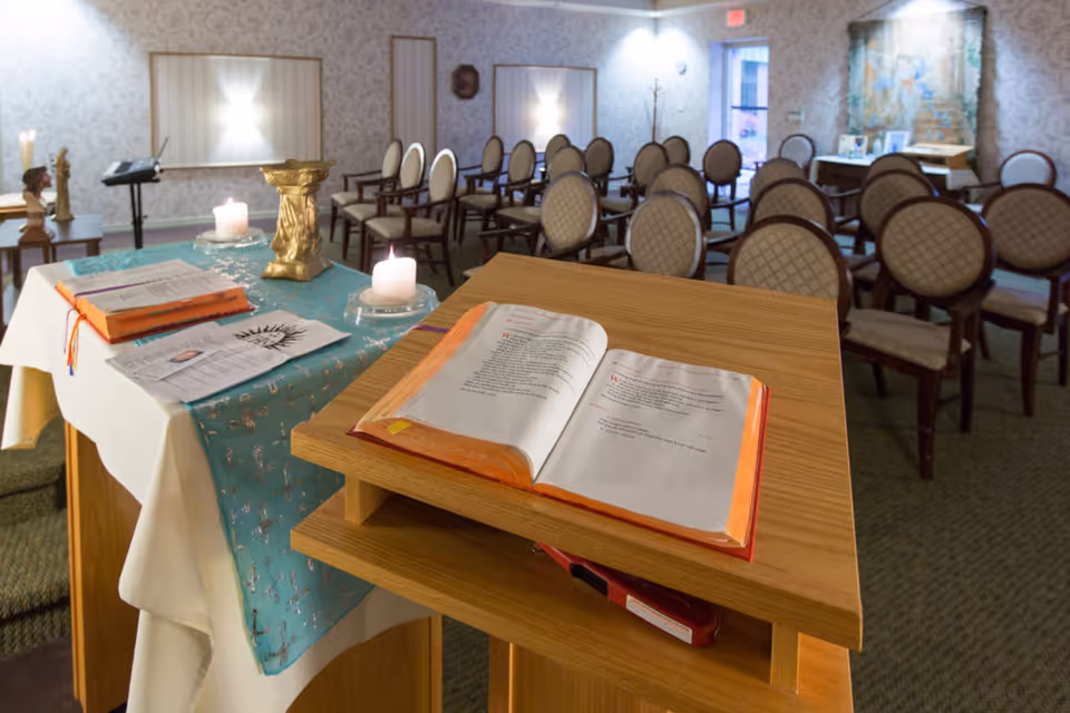 Interior view of a chapel or small worship room with rows of chairs arranged facing a wooden lectern with an open book. A table covered with a white cloth and a blue decorative runner holds candles, religious items, and papers. The room has patterned wallpaper, soft lighting, and a door leading outside.
