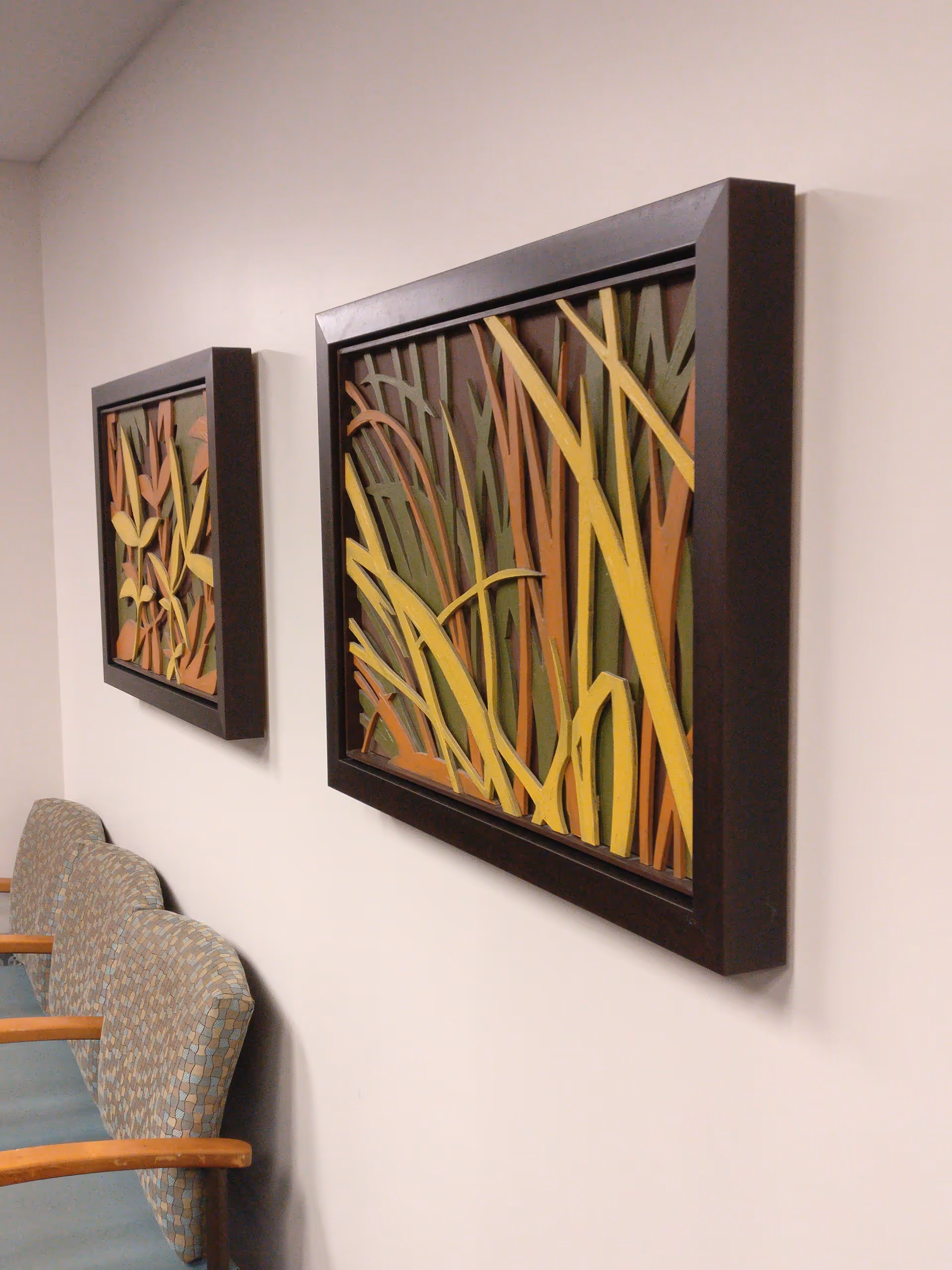Two framed abstract artworks hang on a wall beside patterned chairs in a hallway.