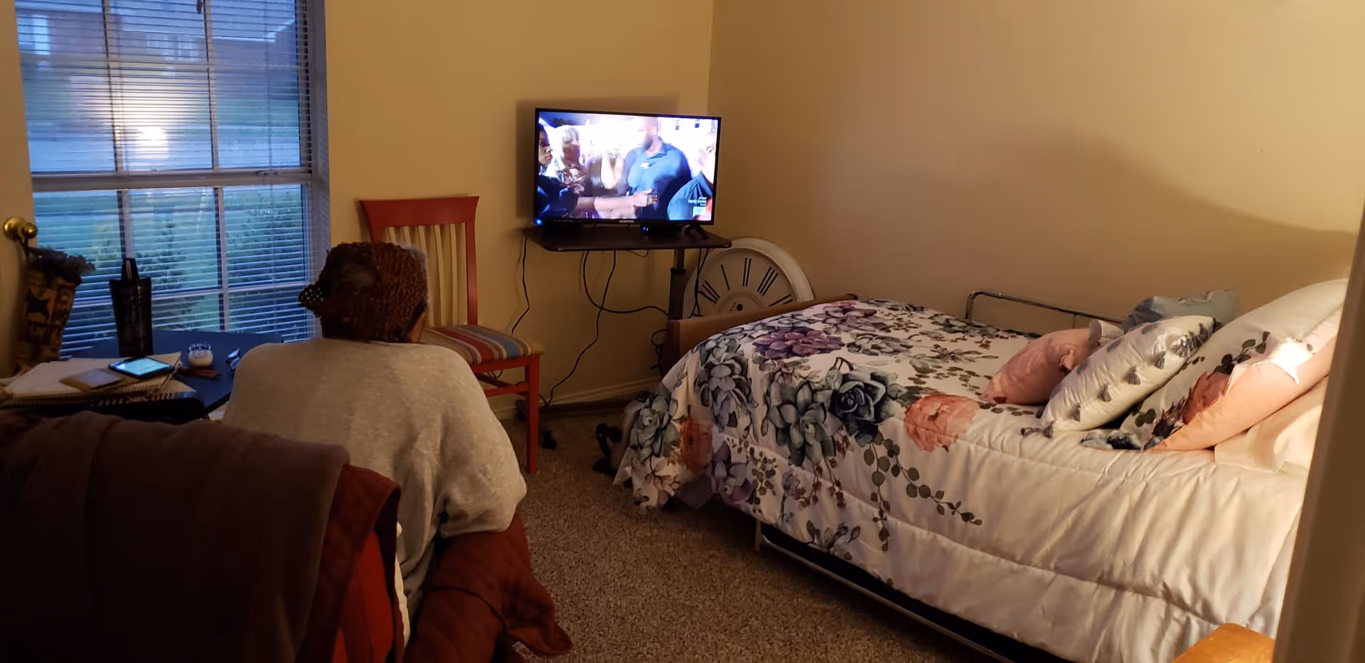 A senior person sitting on a chair facing a television mounted on a small table in a cozy bedroom. The bed has a floral comforter and several pillows. There is a large window with blinds on the left side, a red chair near the TV, and a large decorative clock leaning against the wall behind the TV.