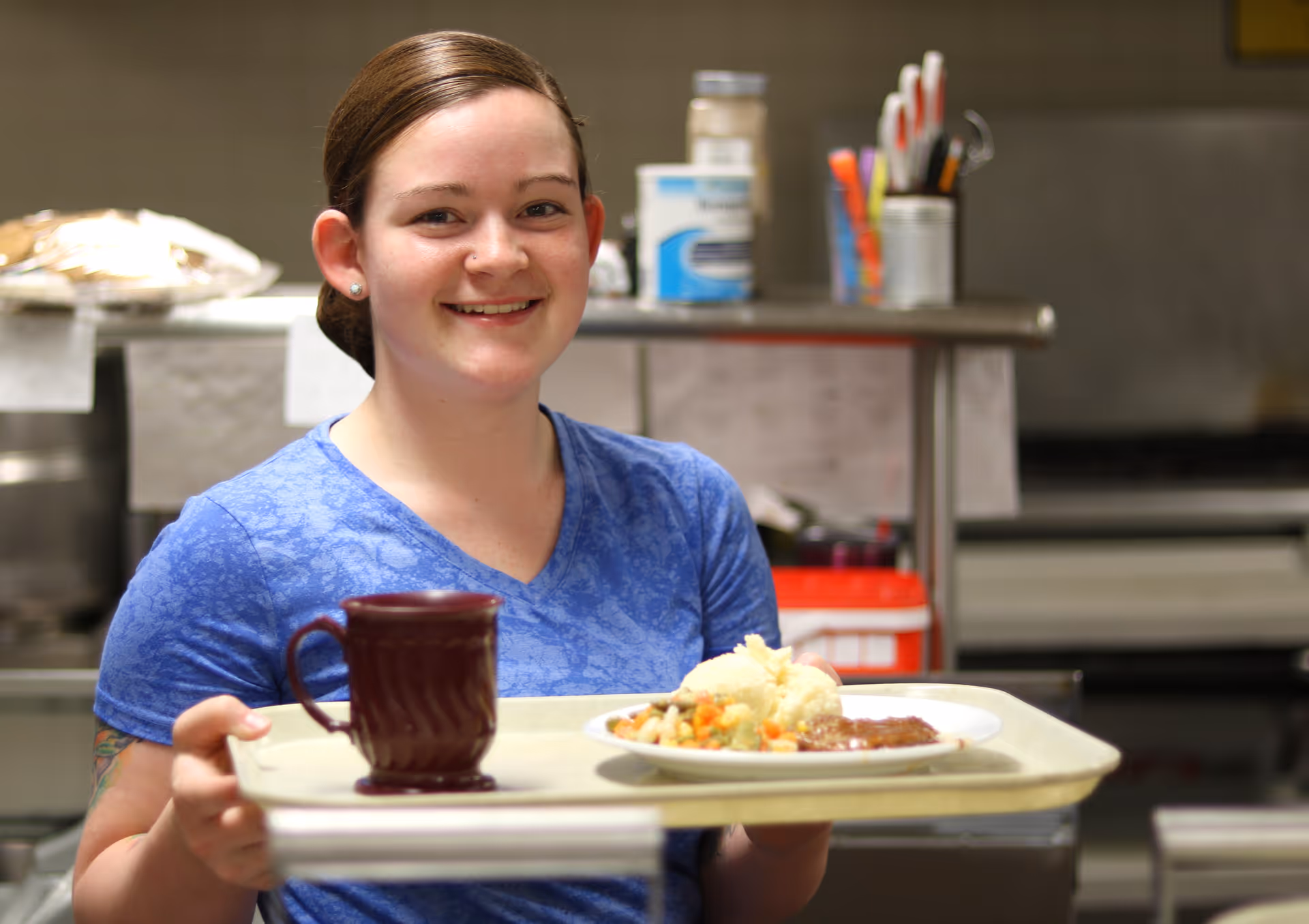 A smiling woman wearing a blue shirt holding a tray with a plate of food and a maroon mug in a kitchen setting.