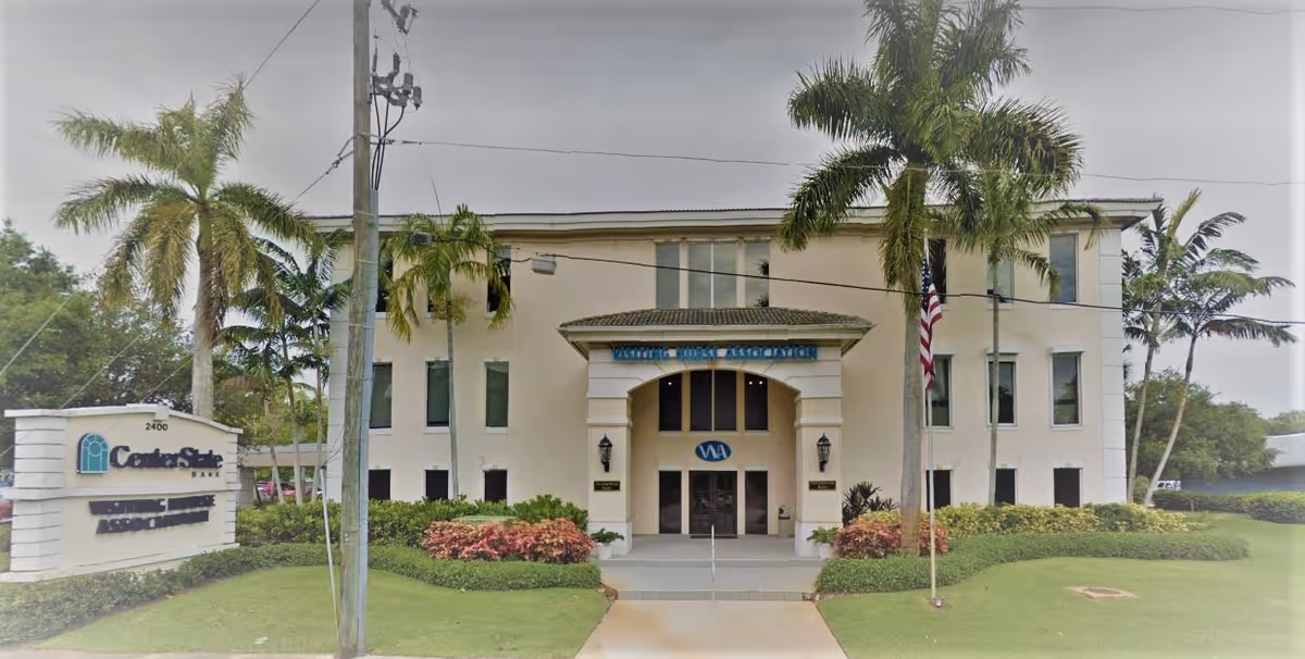 Two-story beige facility with arched entrance, palm trees, and an American flag.