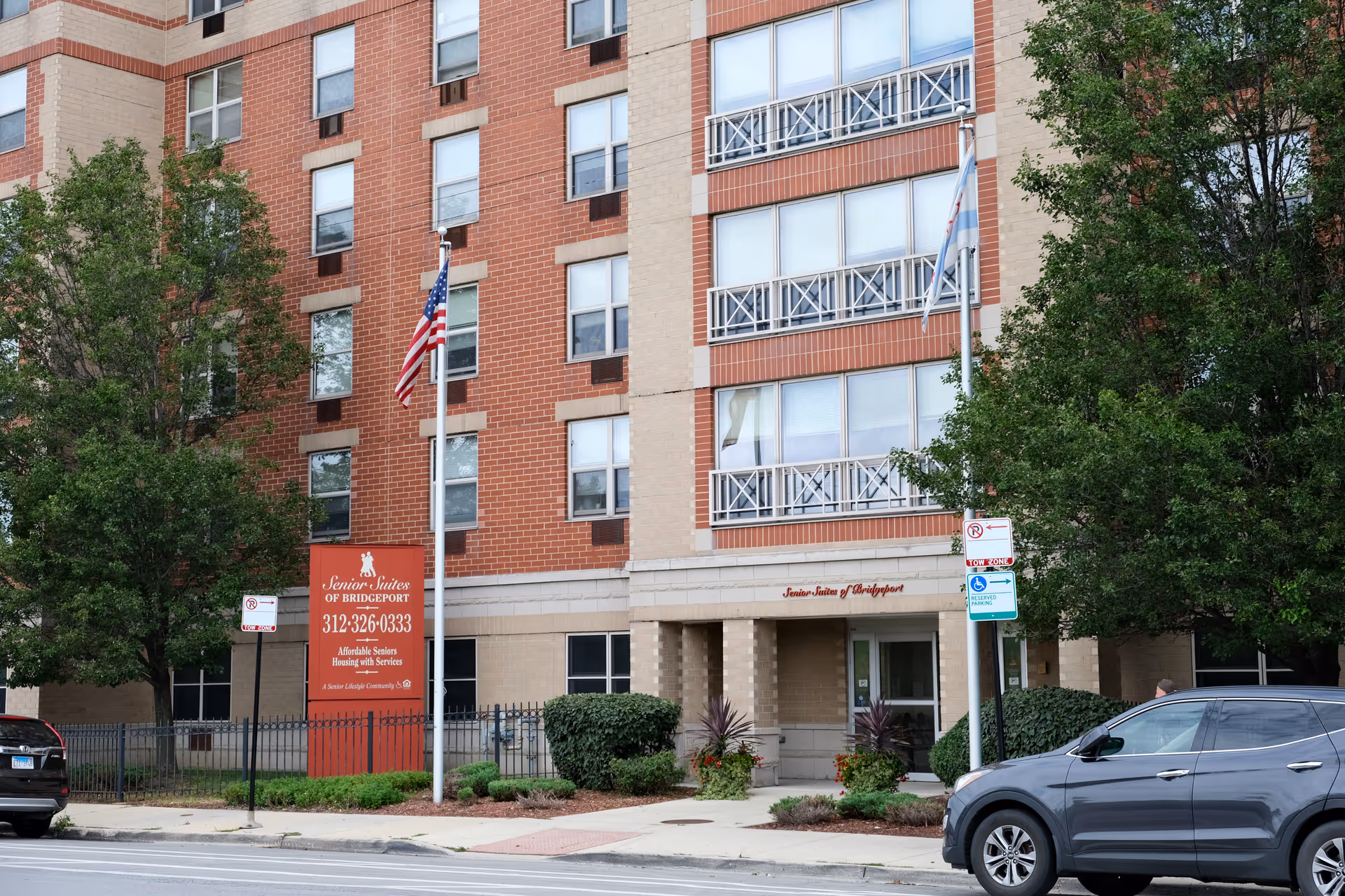 Front exterior of the Senior Suites of Bridgeport building showing the entrance, a red sign with phone number, flagpoles, trees, and parked cars.