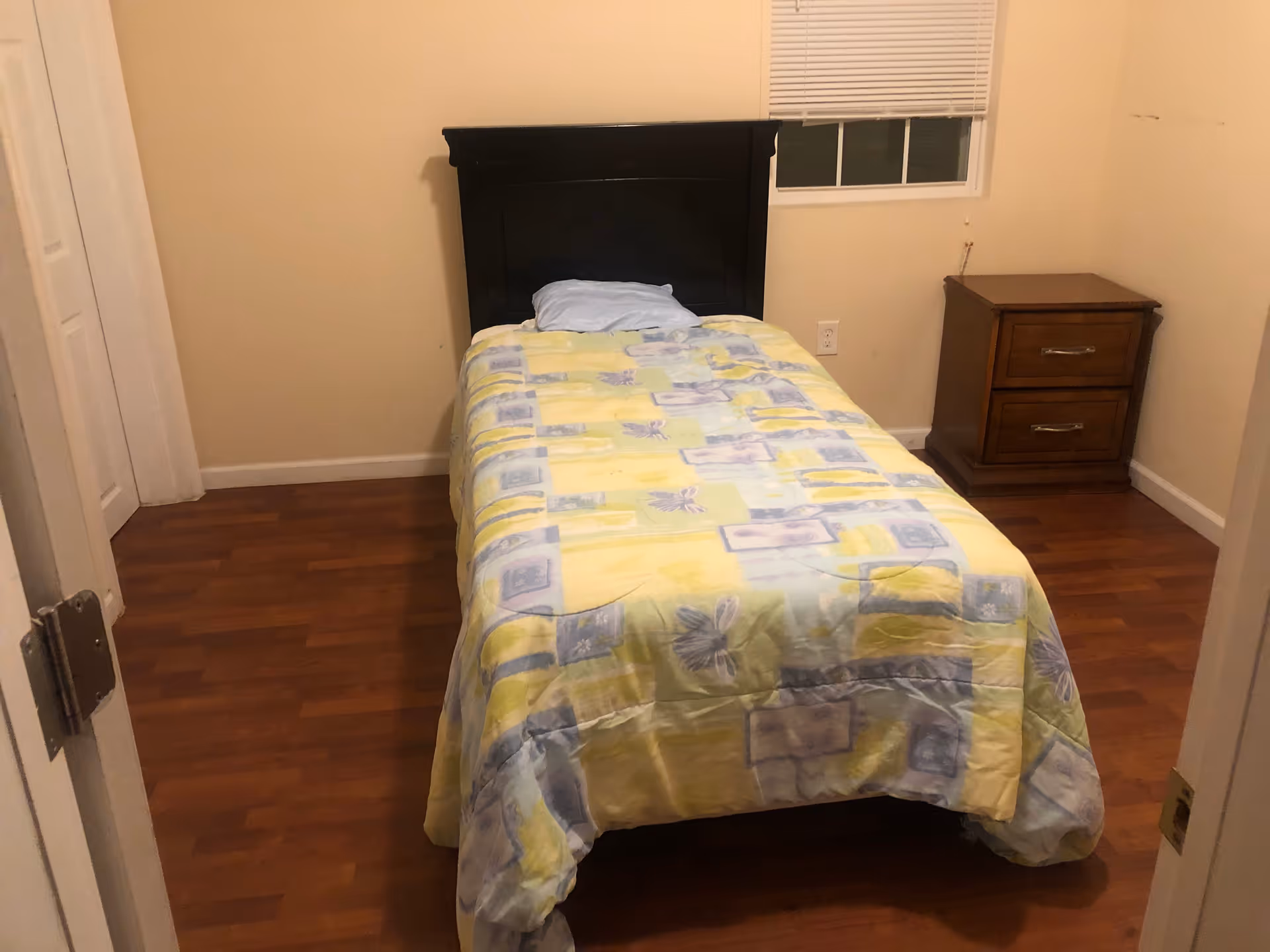 A small bedroom with a single bed covered in a yellow-patterned comforter, a dark headboard, a wooden nightstand, and hardwood floors.