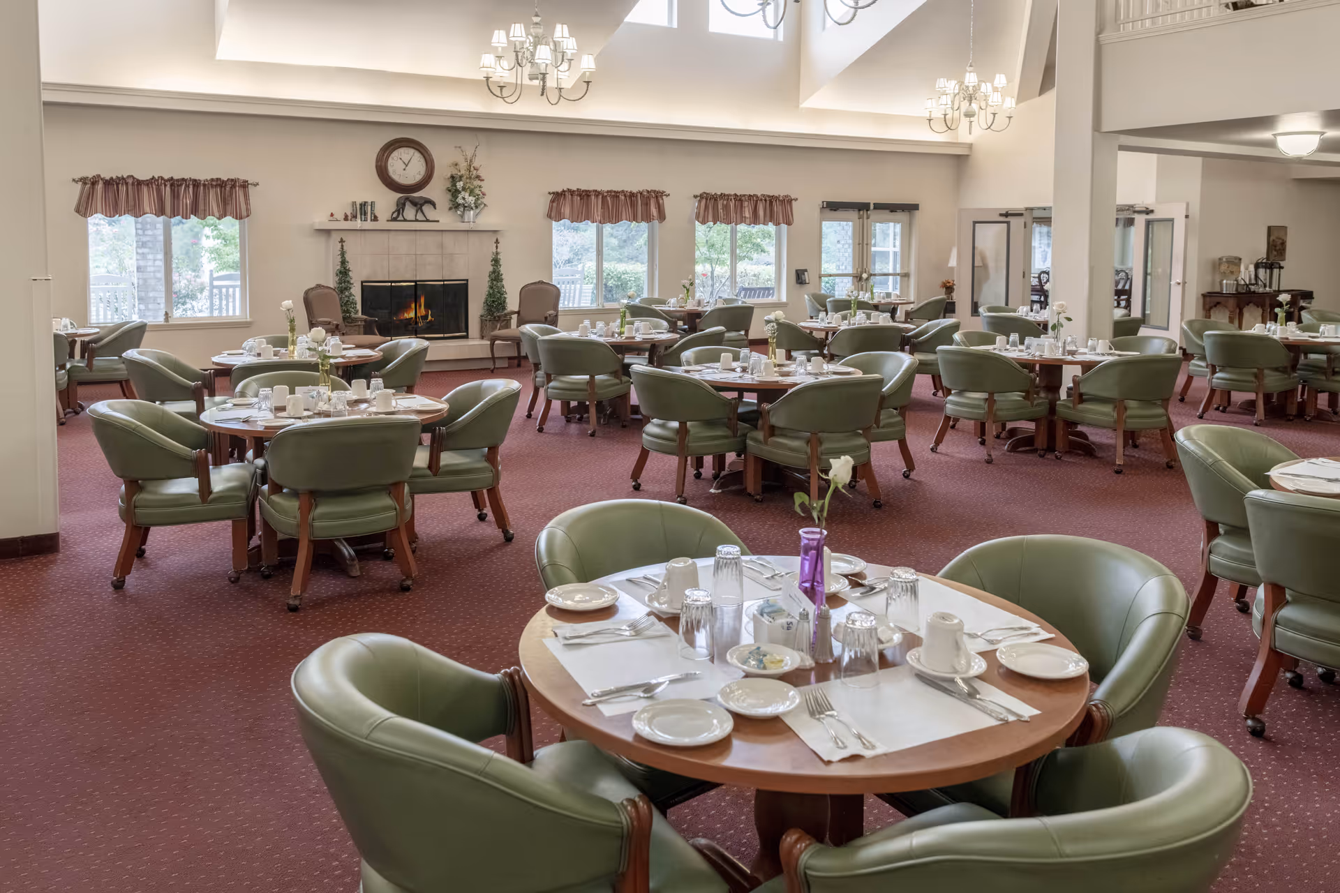 A spacious dining room with multiple round tables set for a meal, each surrounded by green cushioned chairs. The room features a red carpet, large windows with valances, a fireplace with a clock and decorative items above it, and chandeliers hanging from the ceiling.