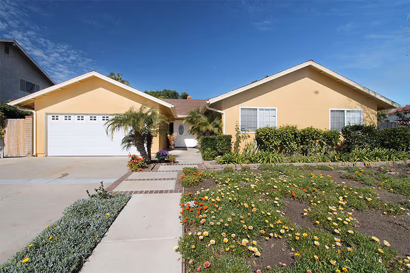 A single-story yellow house with an attached garage and a landscaped front yard with flowering plants and small palm trees under a clear blue sky.