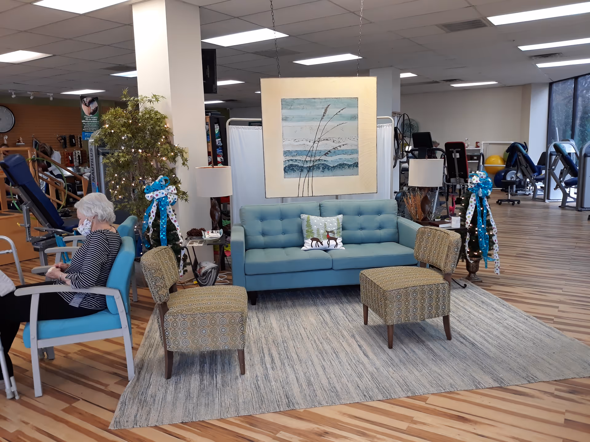 Elderly woman seated left in a bright living lounge with gym equipment visible.