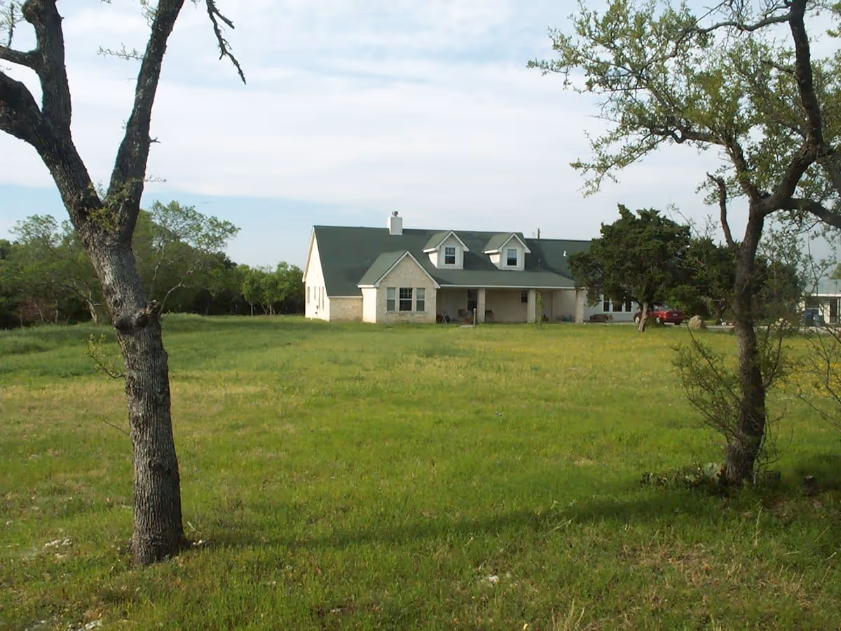 A single-story house with a green roof sits in a large grassy field framed by trees.