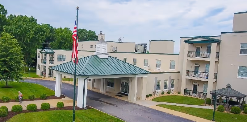 Exterior front entrance of a multi-story retirement community building with a covered porte-cochere, American flag, balconies and landscaped grounds.