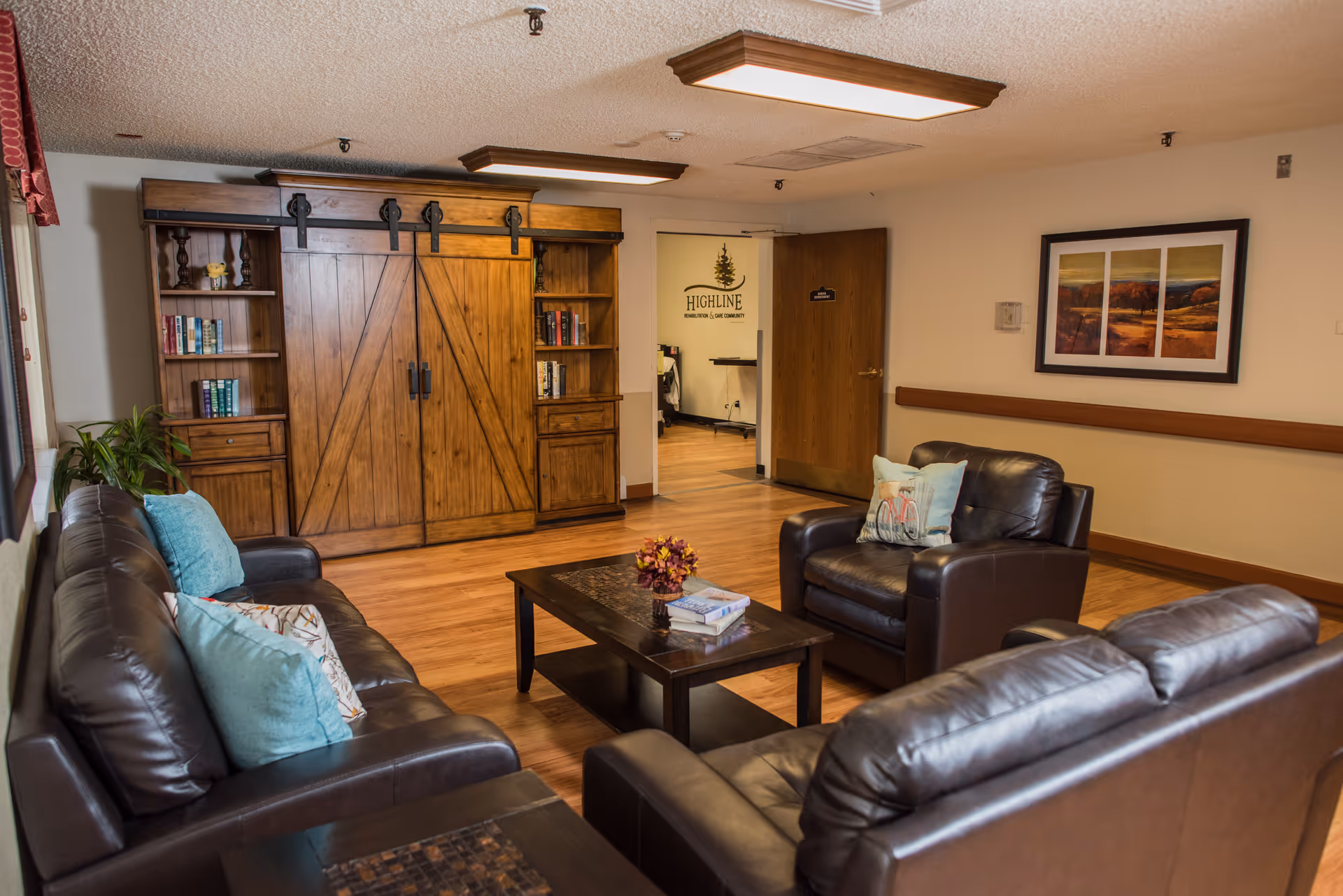 A cozy living room area in a senior living facility with dark brown leather sofas and armchairs arranged around a wooden coffee table. The coffee table has a small flower arrangement and some books on it. There is a large wooden cabinet with sliding barn doors and bookshelves on either side against the wall. A framed landscape painting hangs on the wall above a wooden chair rail. The floor is wood, and the ceiling has recessed lighting. An open door reveals a hallway with a sign that reads 'Highline' on the wall.