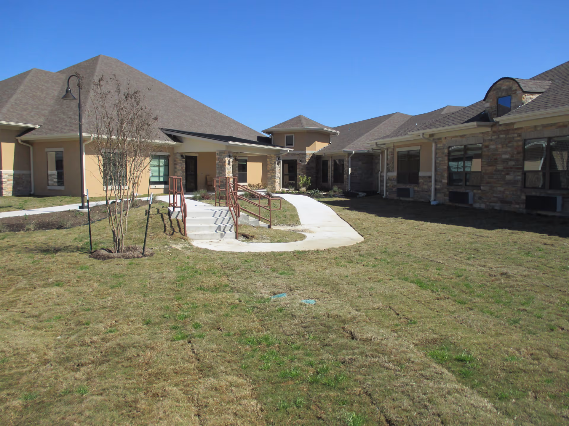 Exterior courtyard and entrance of a single-story senior living facility with a paved walkway, ramps, and a grassy lawn under a clear blue sky.