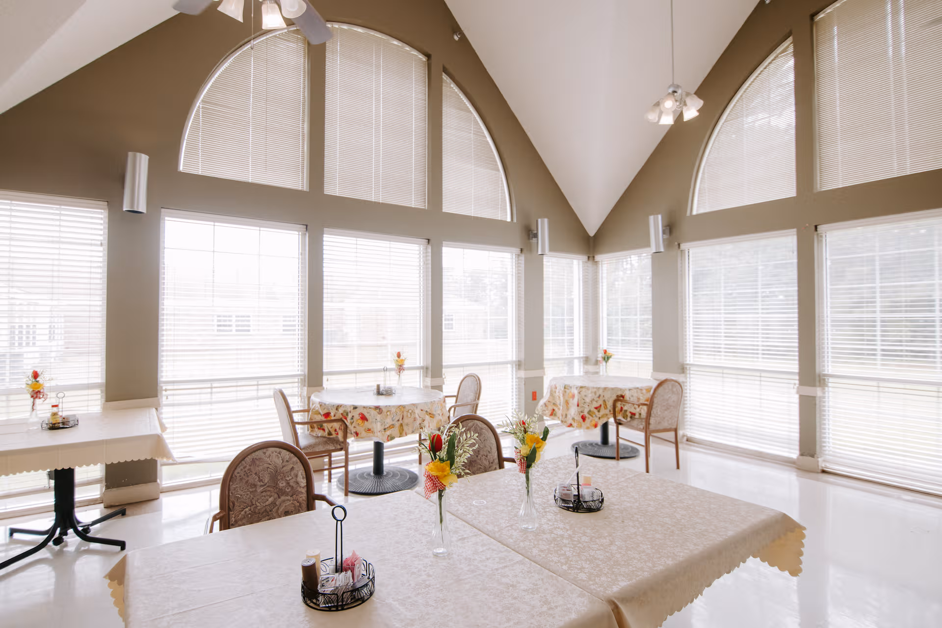 Bright dining room with large arched windows, round tables, chairs, and floral centerpieces.