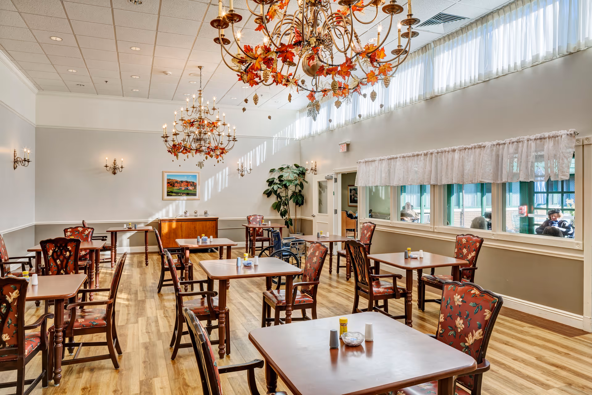 A bright dining room with wooden floors and multiple square wooden tables surrounded by floral upholstered chairs. The room features large windows with sheer curtains, decorative chandeliers adorned with autumn leaves, and wall sconces providing additional lighting. A wheelchair is visible near one of the tables, and a few people can be seen through the windows in an adjacent area.