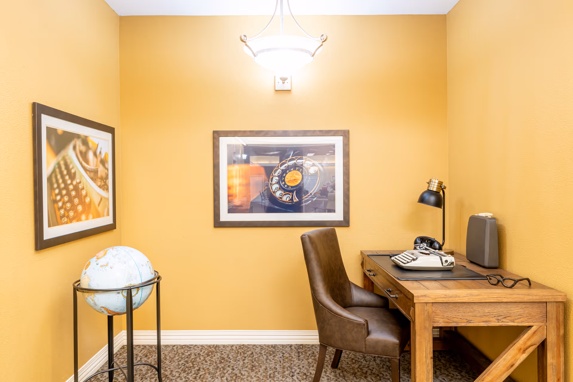 Small office nook with a wooden desk and leather chair, a vintage typewriter and lamp on the desk, a globe on a stand, and framed pictures on yellow walls.