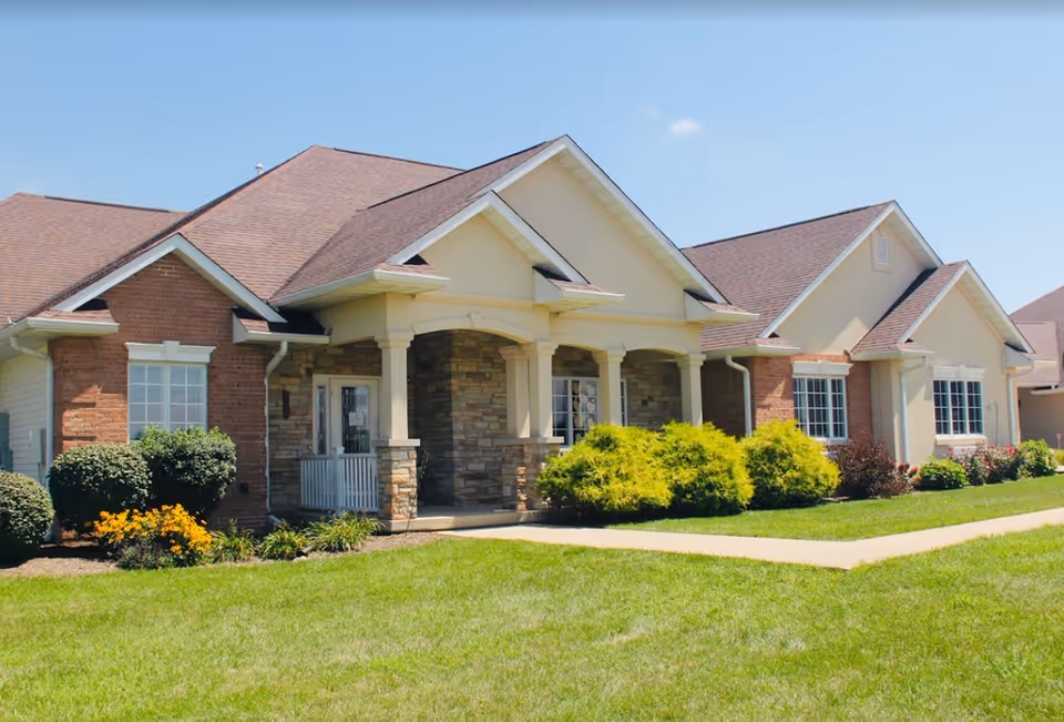Exterior view of a single-story senior living facility building with a combination of brick and stone facade, a covered entrance supported by columns, well-maintained green lawn, shrubs, and flowers under a clear blue sky.