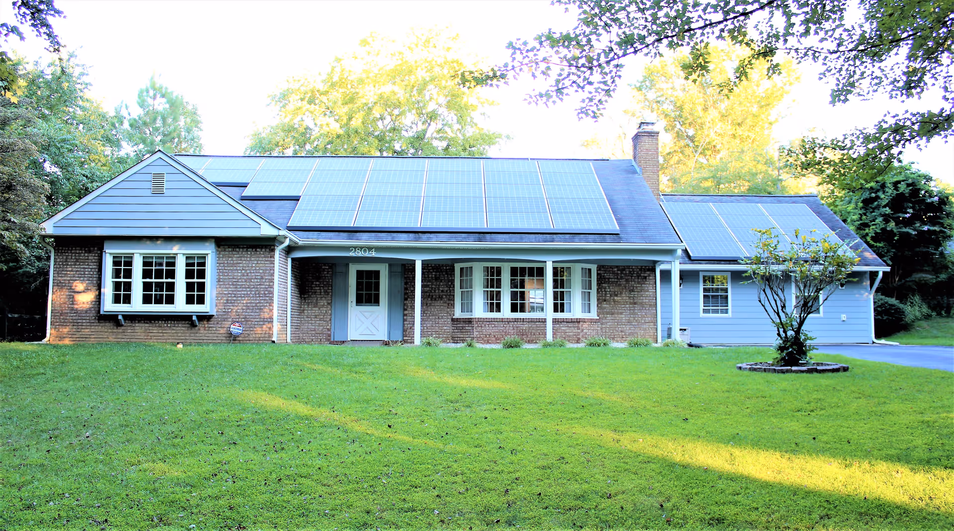Single-story brick and siding house with solar panels on the roof, a covered front porch, and a large grassy lawn.
