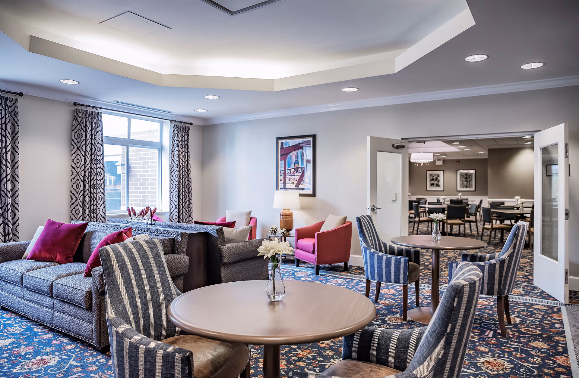 A well-lit common area in a senior living facility featuring a seating arrangement with patterned sofas and armchairs, round wooden tables with small flower vases, and a carpet with a floral design. The room has large windows with patterned curtains, recessed ceiling lights, and a framed artwork on the wall. Double doors open to an adjoining dining area with multiple tables and chairs.