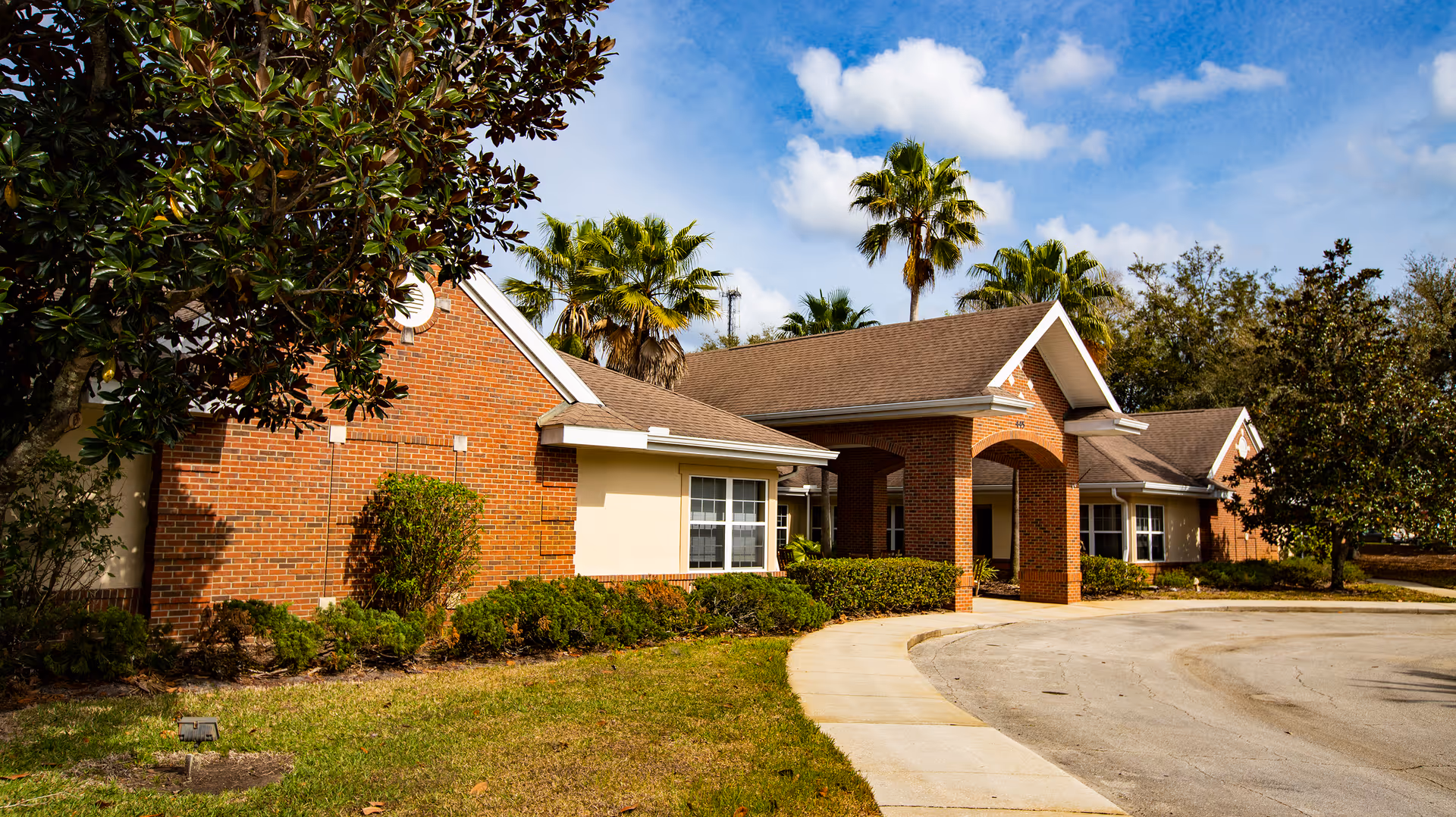 Brick senior living facility with arched entrance, curved driveway, and palm trees