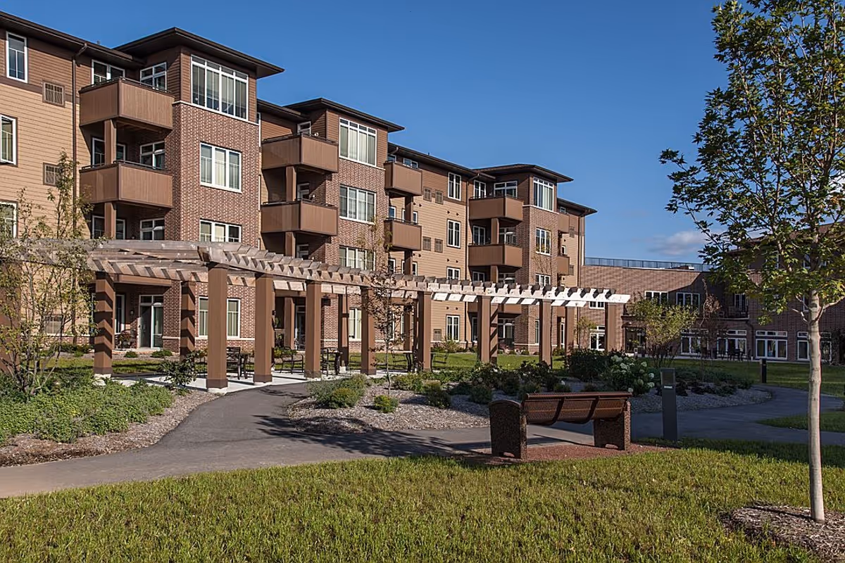 Outdoor view of a senior living facility named Dickson Hollow featuring a multi-story building with balconies, a pergola structure, walking paths, landscaped greenery, trees, and a bench under clear blue sky.
