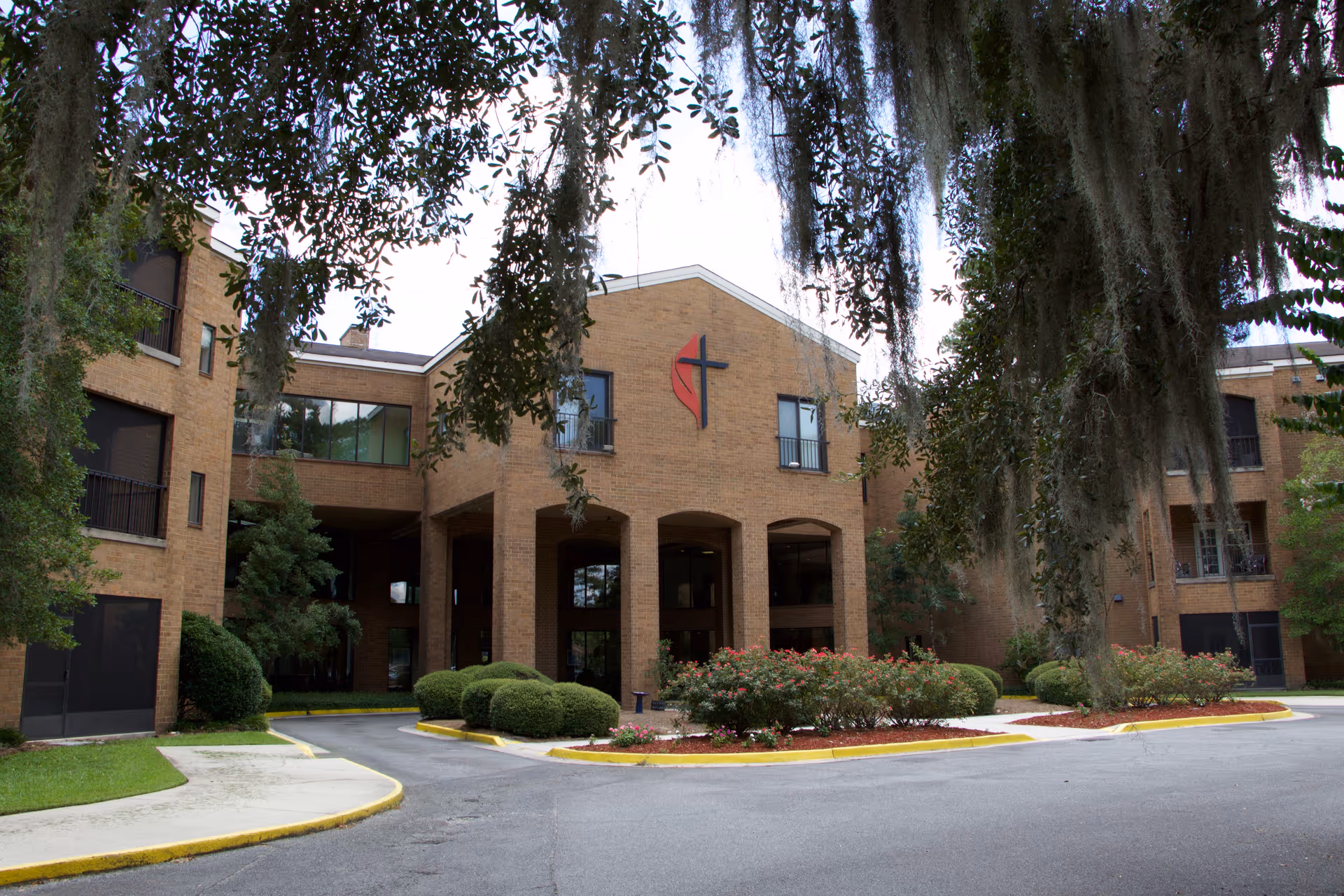 Exterior view of a brick senior living facility building with a covered entrance supported by columns. The building has multiple windows and is surrounded by landscaped bushes and trees with hanging Spanish moss.