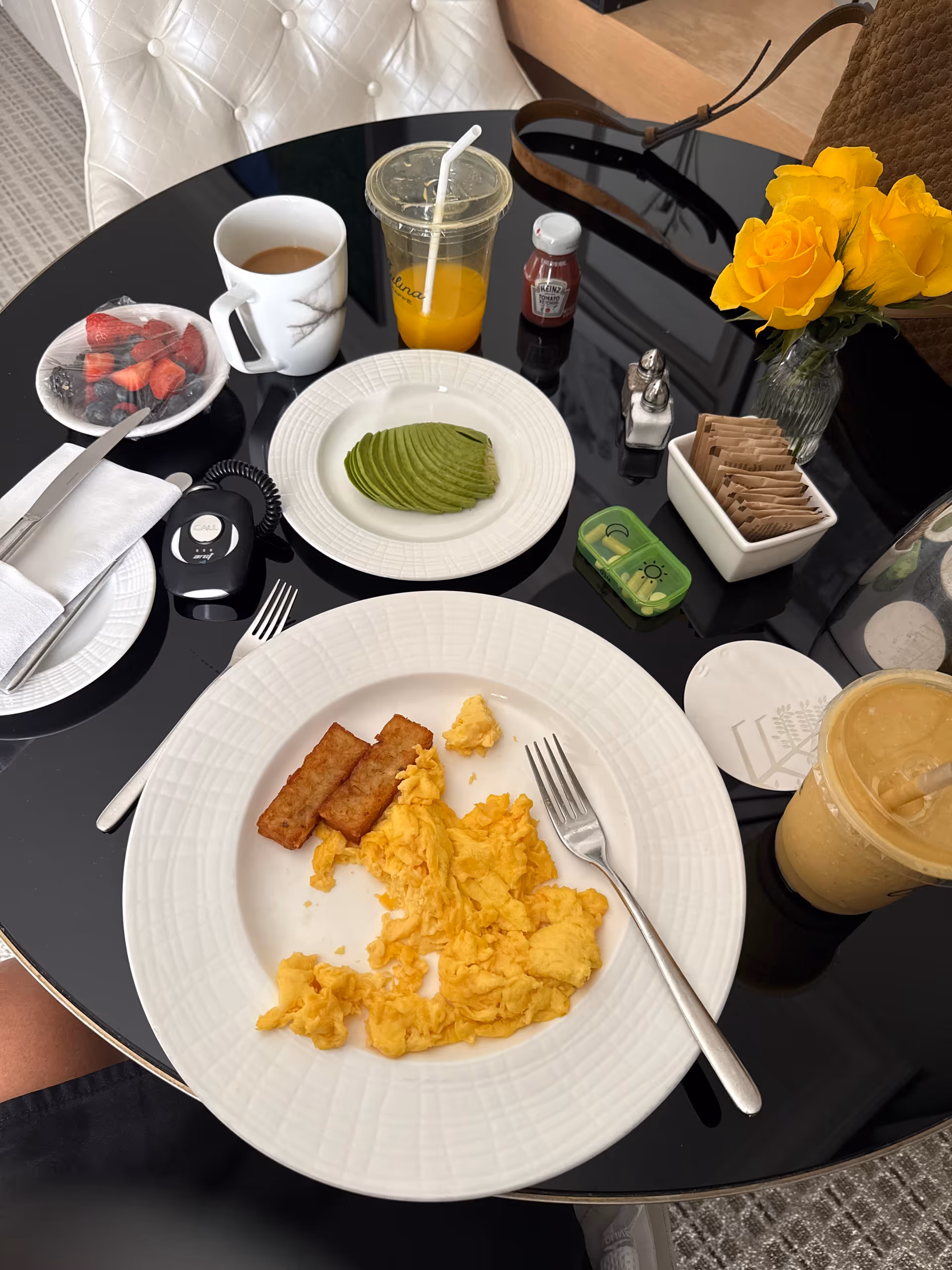 Breakfast on a glossy dining table with eggs, avocado slices, toast, coffee, juice, and flowers.