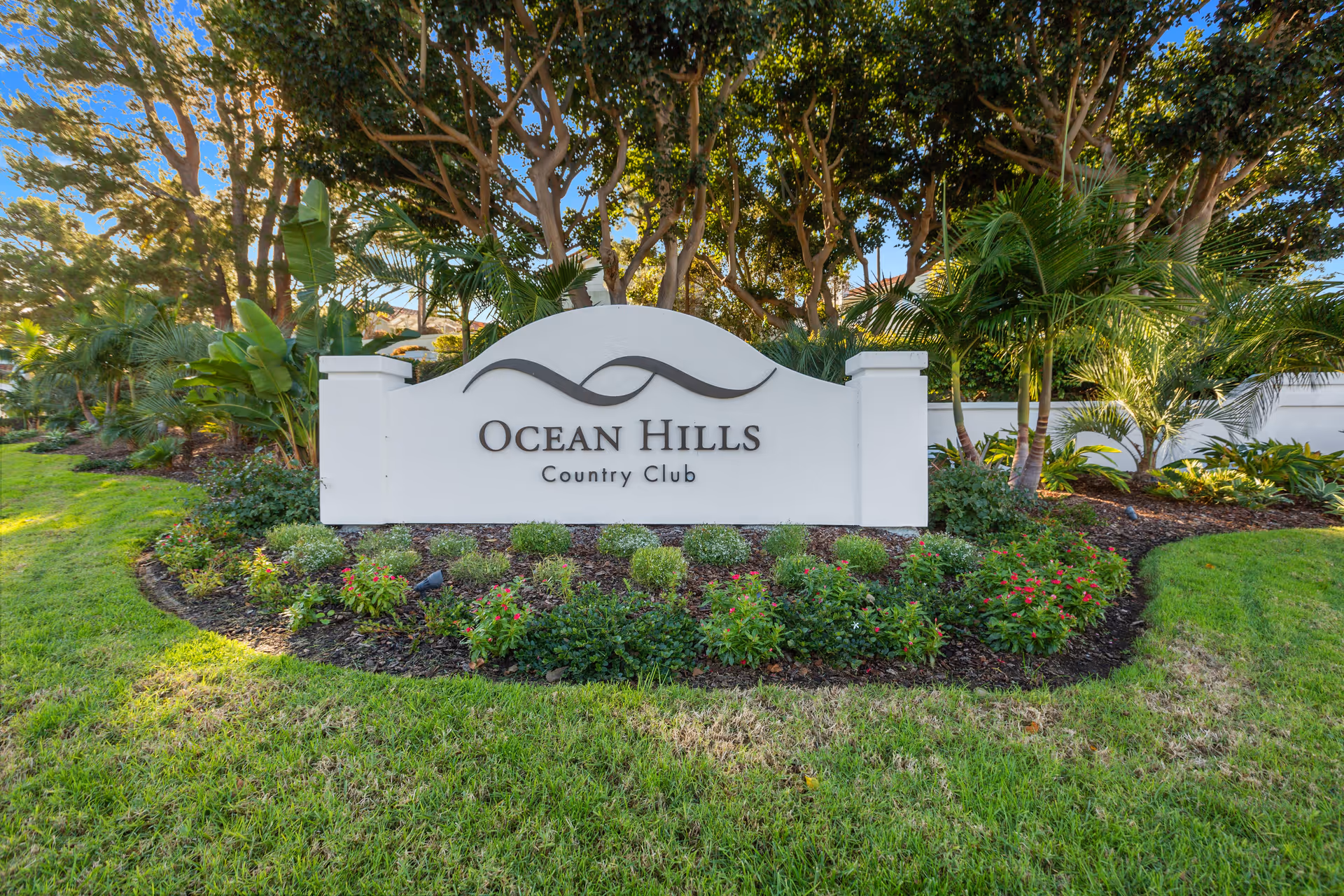 White entrance sign reading "Ocean Hills Country Club" surrounded by landscaped plants and trees.
