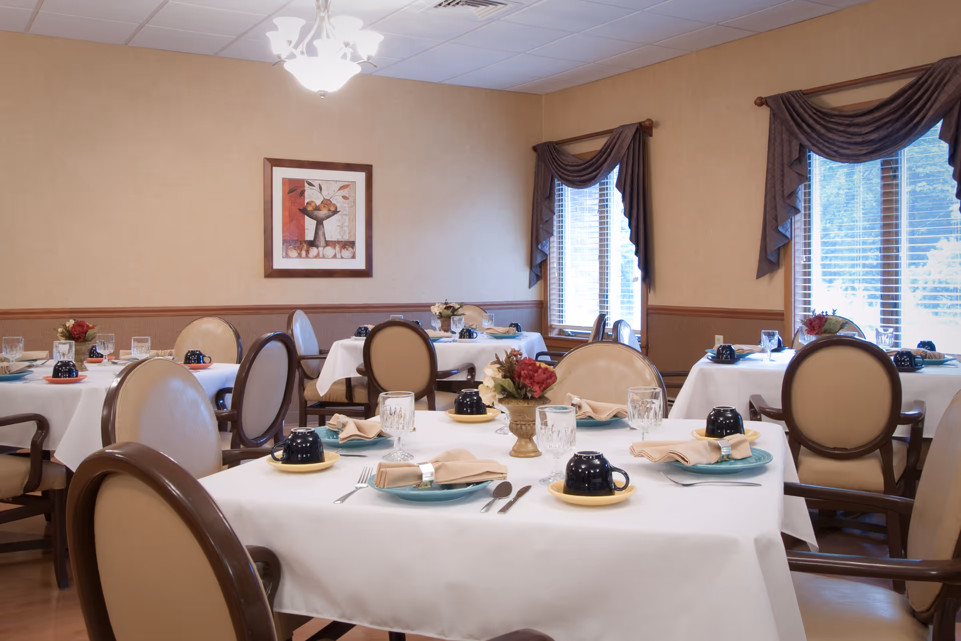 A dining room with neatly set tables, upholstered chairs, and floral centerpieces by windows.