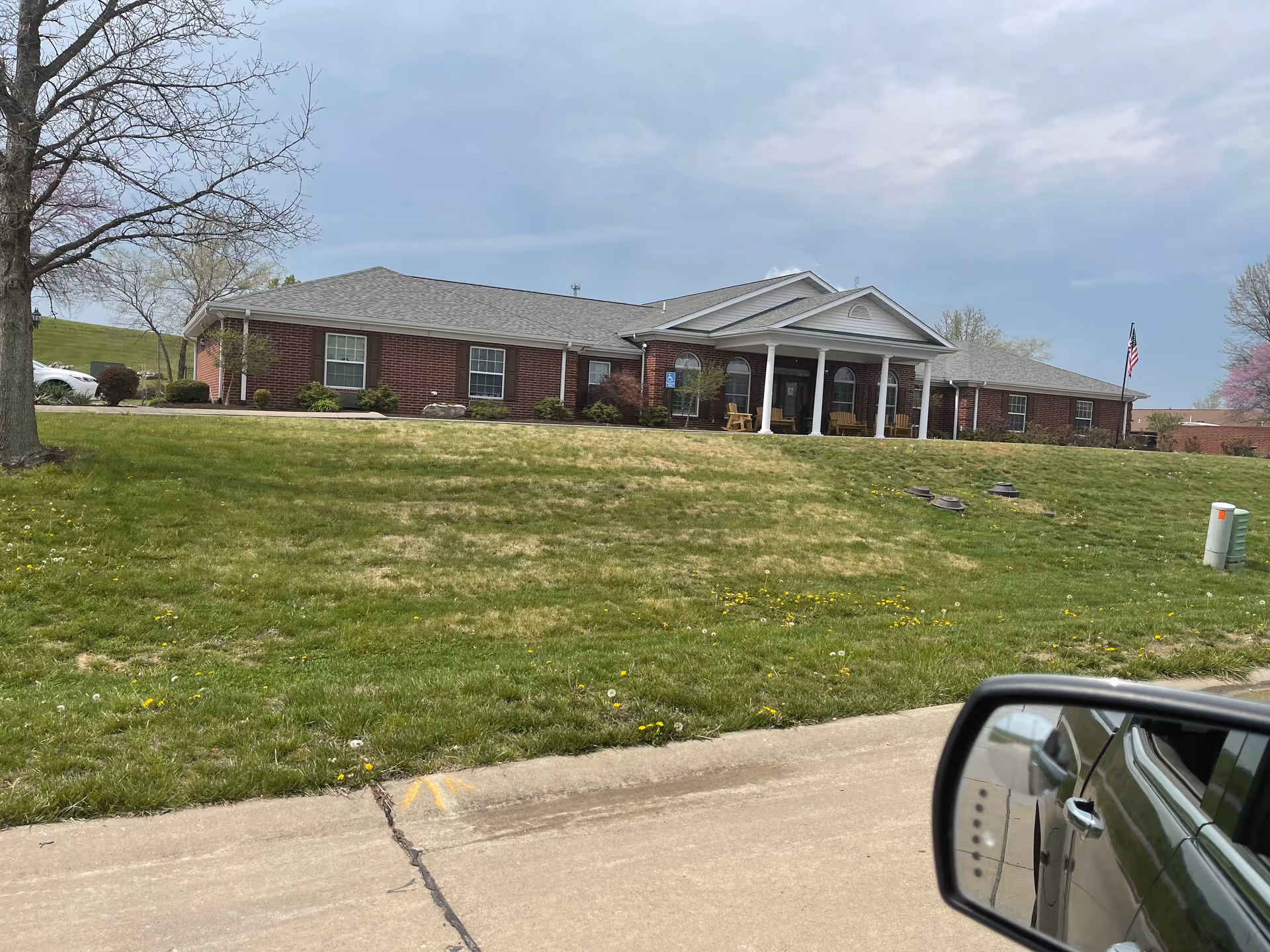 Brick senior living facility with white columns, a front porch, manicured lawn, and American flag.