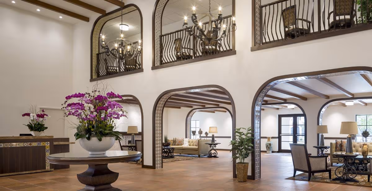 Spacious and elegant senior living facility lobby with high ceilings, decorative arches, tiled floor, a round wooden table with a large floral arrangement, seating areas with chairs and lamps, and a reception desk in the background.