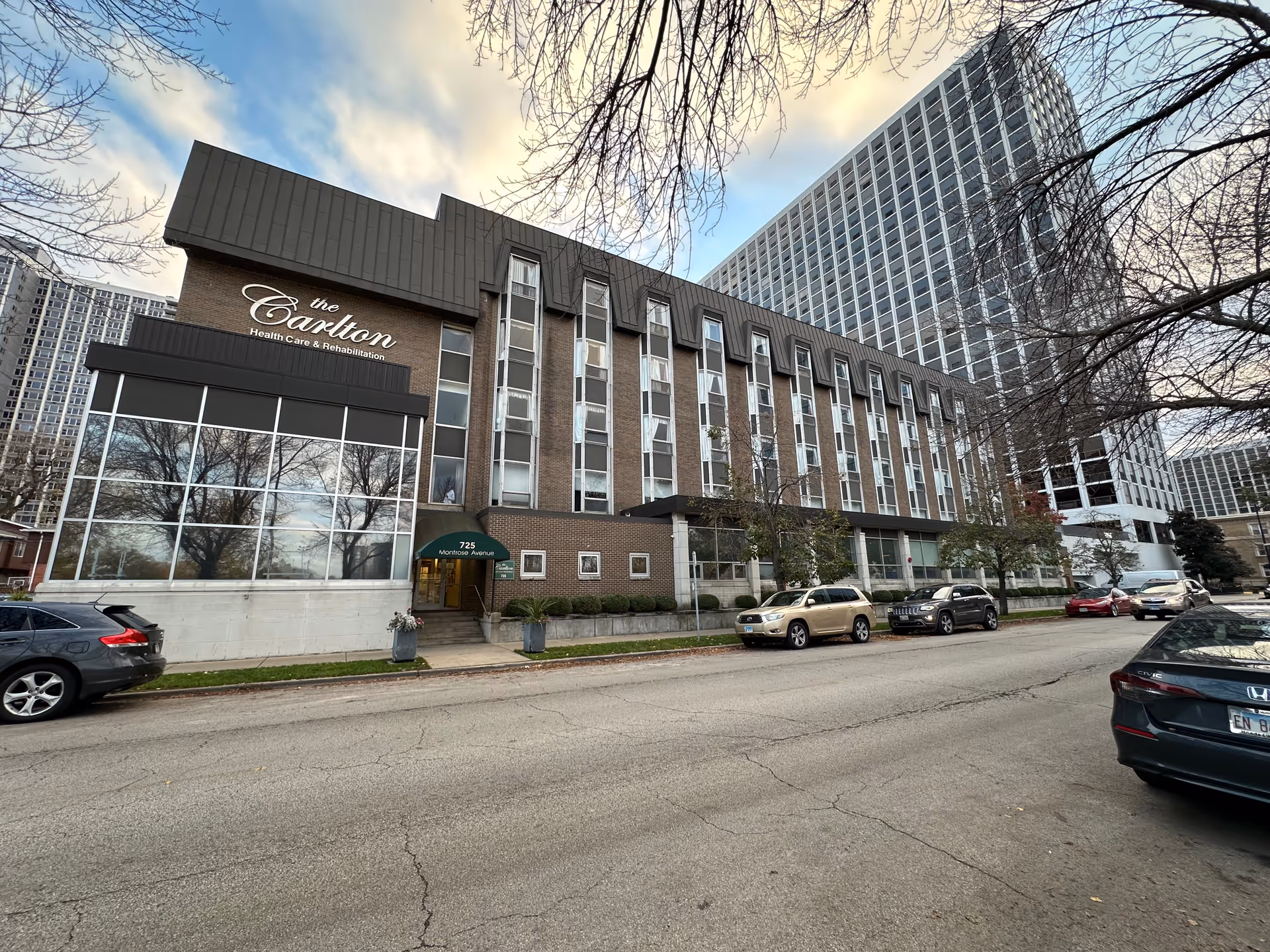 Exterior view of The Carlton Health Care & Rehabilitation building, a multi-story brick structure with large windows and an entrance canopy labeled 725 Montrose Avenue. Several cars are parked along the street in front of the building, and leafless tree branches frame the top of the image under a partly cloudy sky.