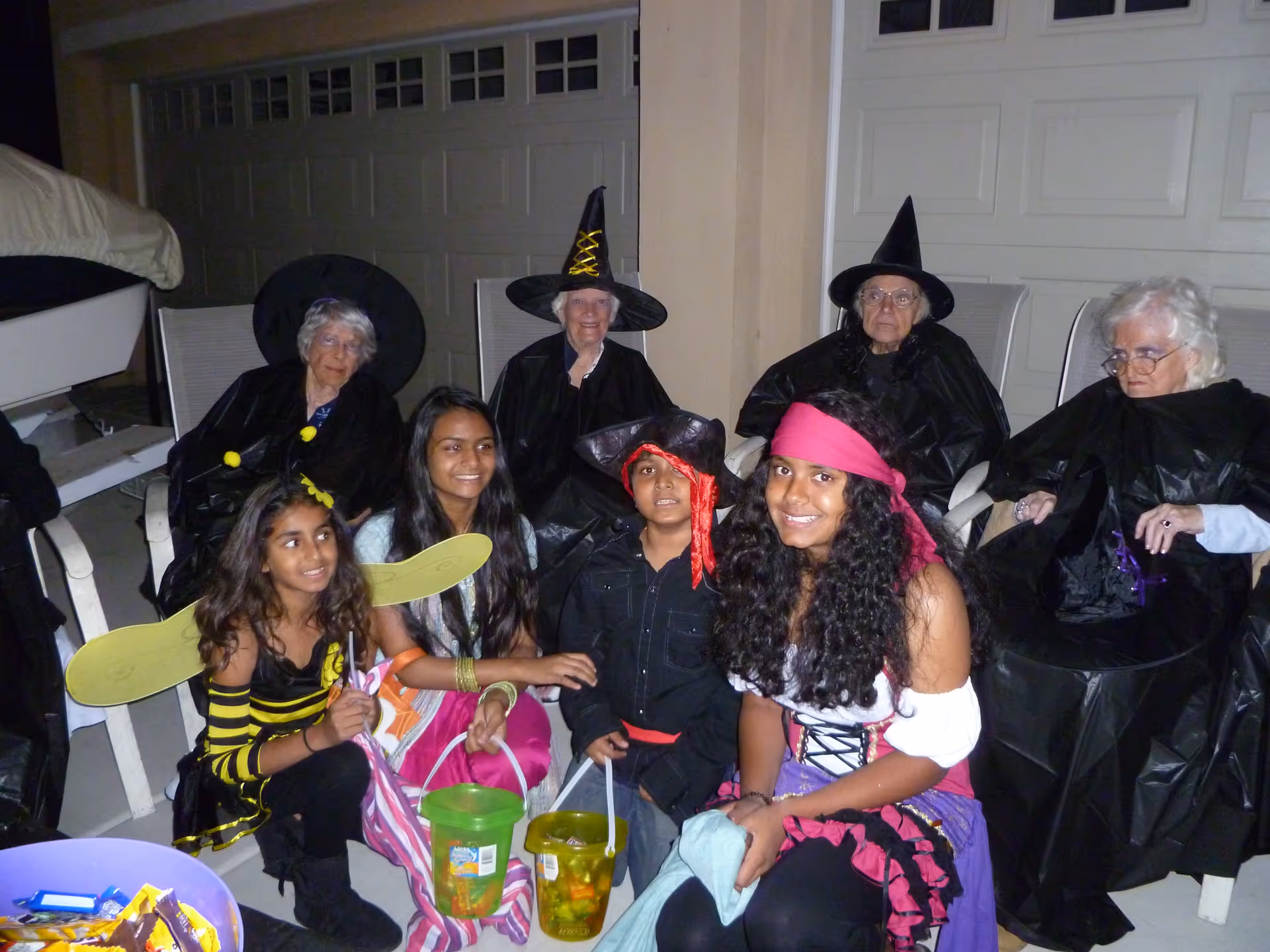 A group of elderly women dressed as witches sitting on chairs outside, accompanied by four children in Halloween costumes including a bee, a pirate, and other festive outfits, holding buckets for trick-or-treating.
