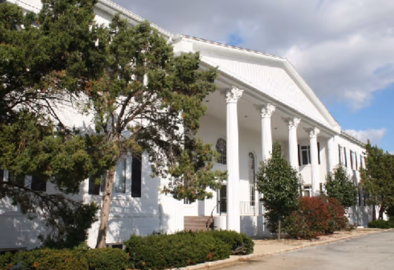 White building with large pillars and a manicured front yard.