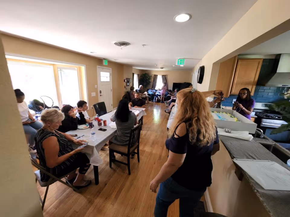 Residents and staff gather around a long dining table in a sunny senior living area.