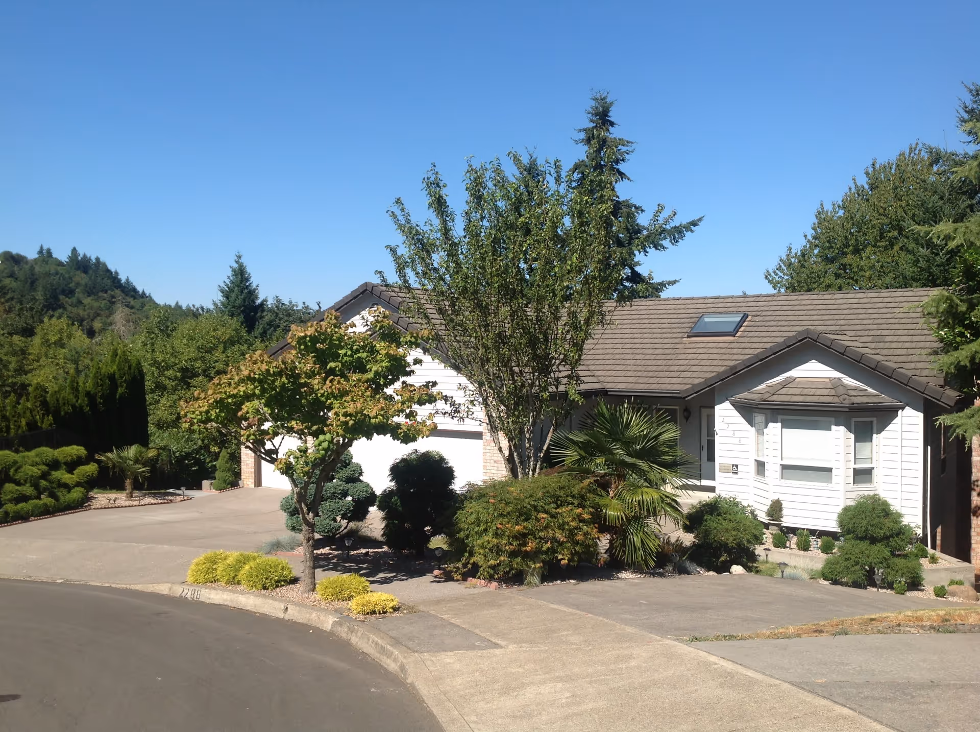 Suburban single-story house with a landscaped front yard, driveway, and trees under a clear blue sky.