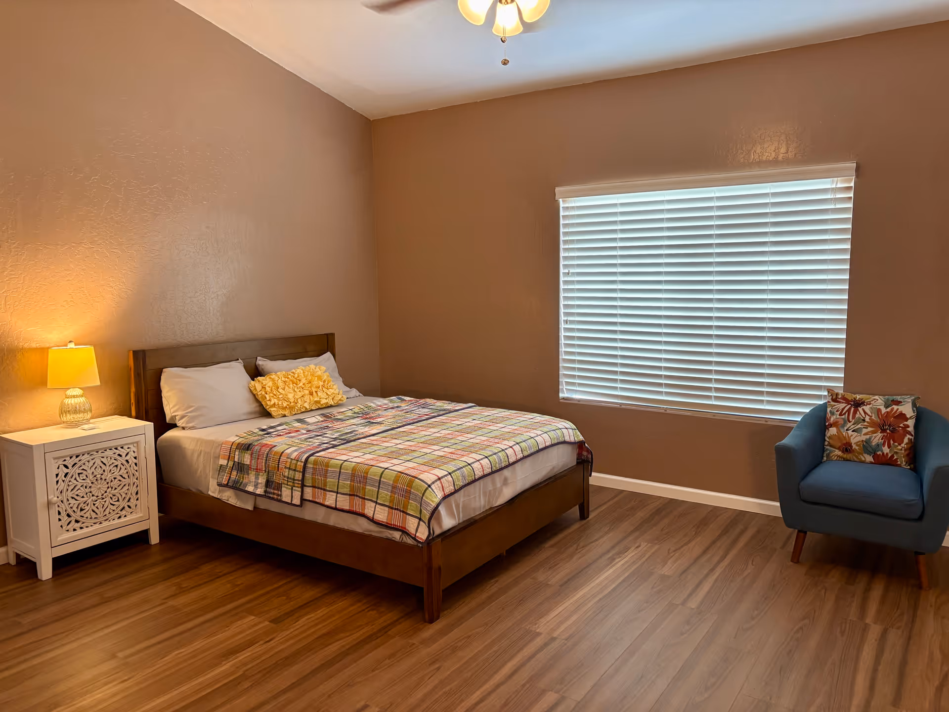 A cozy bedroom with a bed, nightstand, lamp, and blue armchair by a window.