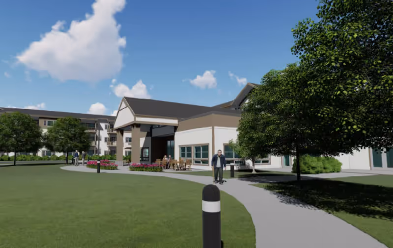 Outdoor view of a senior living facility with a paved walkway, green lawn, trees, and a building with a covered entrance. Several people are walking or sitting outside near the entrance under a clear blue sky with some clouds.