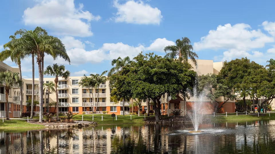 View of a senior living facility with a pond and a water fountain in the foreground, surrounded by palm trees and other greenery, with a multi-story building in the background under a partly cloudy sky.