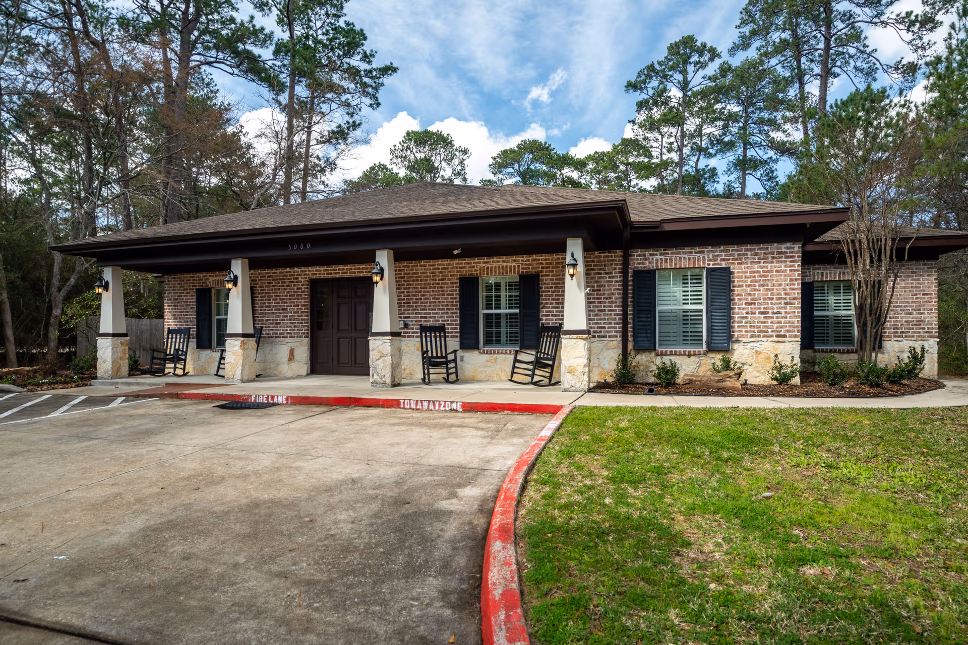 Single-story brick senior living facility exterior with a covered front entrance and rocking chairs.