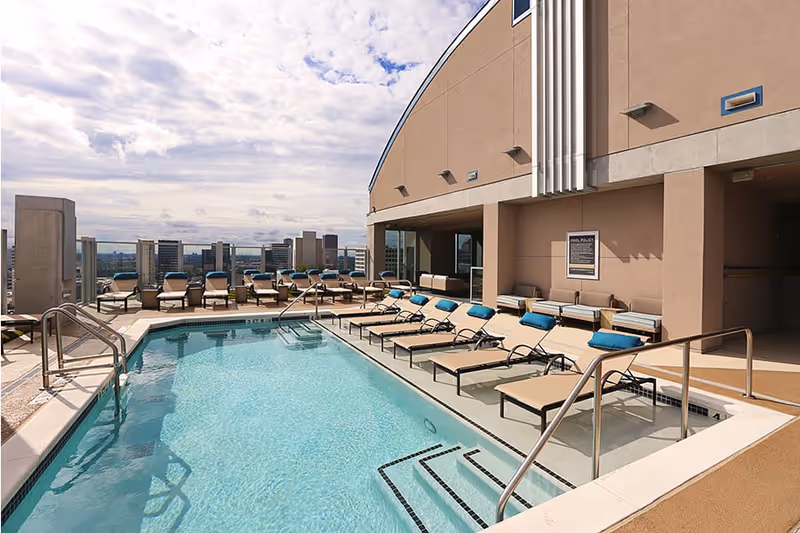 Outdoor rooftop swimming pool with clear blue water, surrounded by beige lounge chairs with blue cushions. The pool area is adjacent to a building with a tan exterior wall and a sign displaying pool rules. In the background, a city skyline is visible under a partly cloudy sky.