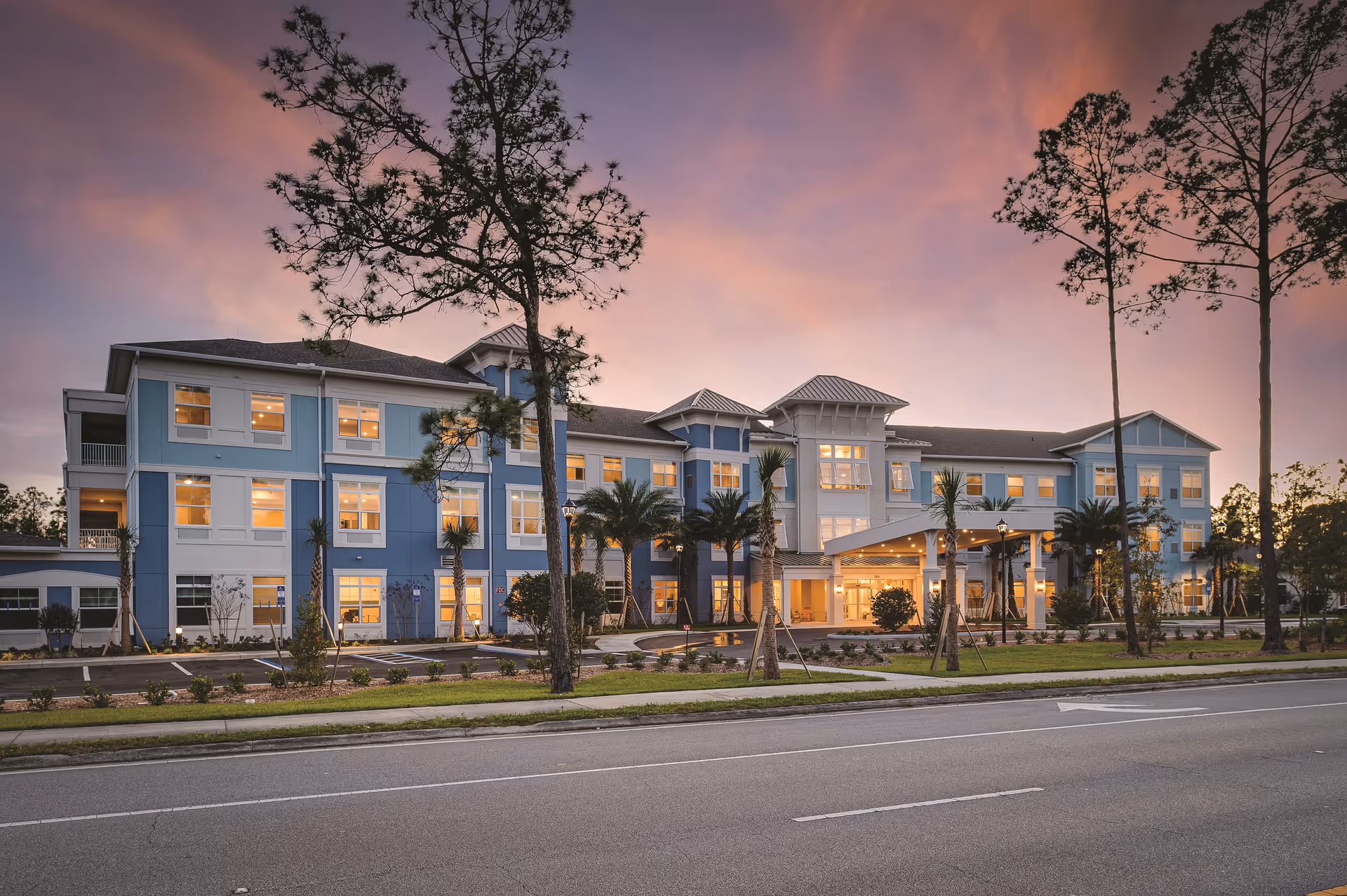 Exterior view of a three-story senior living facility building named Sancerre at Palm Coast during sunset, with warm lights glowing from the windows, palm trees and other landscaping in front, and a clear sky with pink and purple hues.