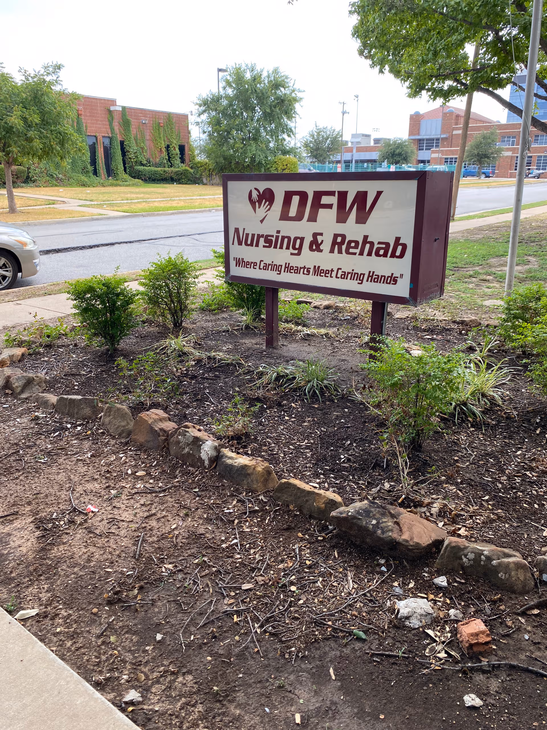 Exterior view of a DFW Nursing & Rehab sign with landscaping and a street behind.