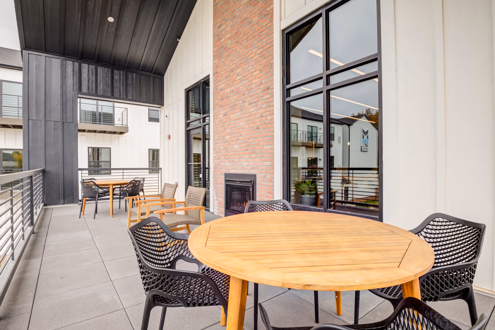 Outdoor balcony area with round wooden tables and black chairs, a brick wall with a built-in fireplace, large windows reflecting the building exterior, and a metal railing along the edge of the balcony.
