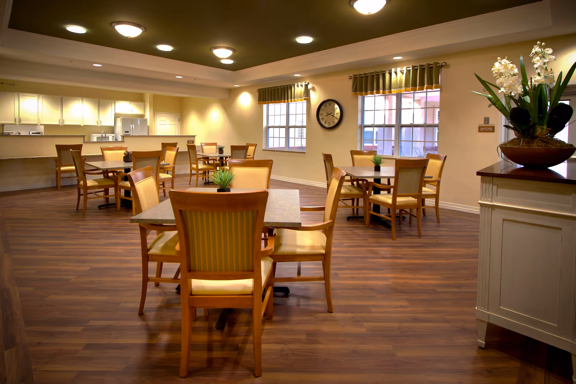 A spacious dining room with several tables and chairs arranged neatly on a wooden floor. The room has large windows with green curtains, a wall clock, and a kitchen area with white cabinets and appliances in the background. There are small plants on the tables and a large floral arrangement on a white cabinet to the right.