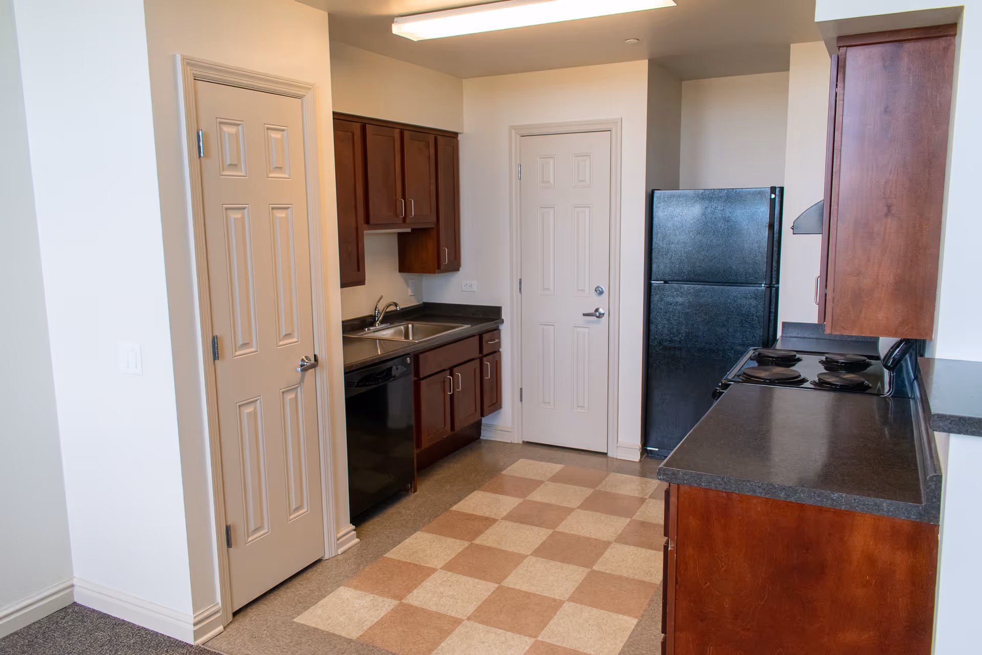 Compact kitchen with dark wood cabinets, fridge, stove, sink, and tiled checkered floor.
