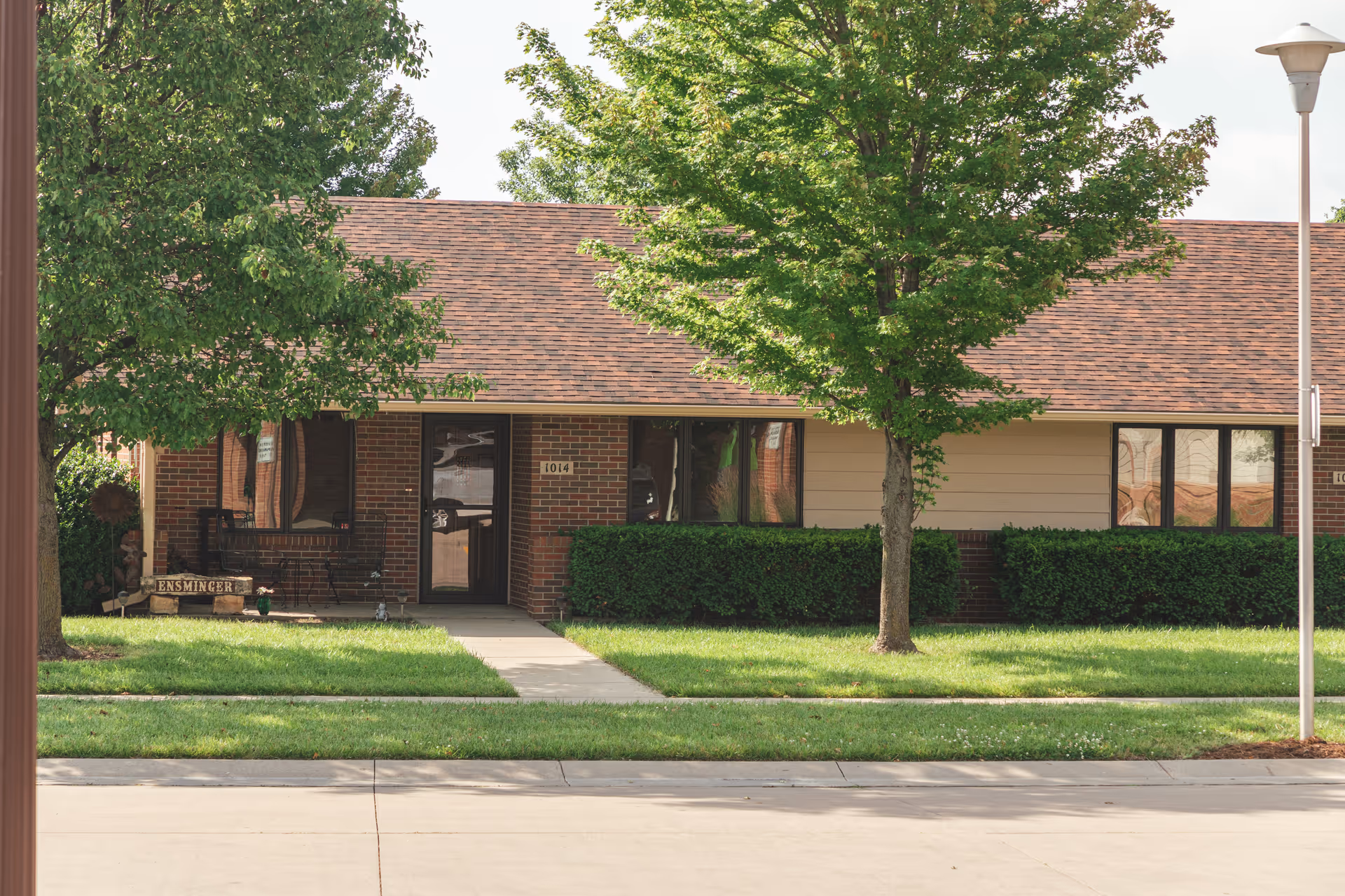 Exterior view of a single-story brick and siding building with a brown shingled roof, green bushes, and two trees in front. A concrete walkway leads to a black door with the number 1014 above it. There is a streetlamp on the right side and a small porch area with a bench and a sign that reads 'ENSMINGER'.