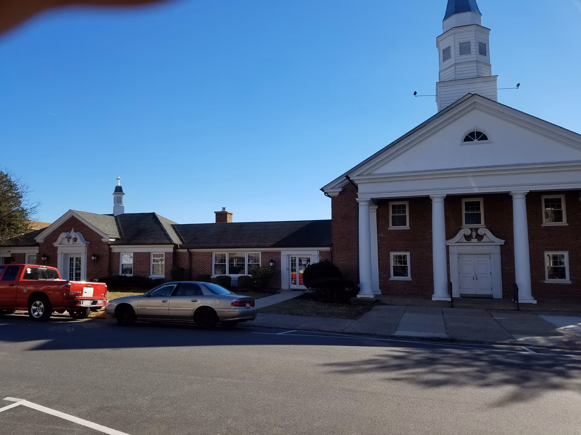 Brick facility with white columns and a tall steeple; cars parked along the street.