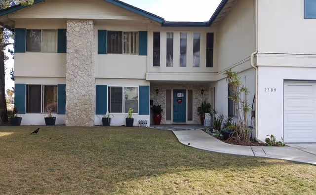 Two-story senior living facility front exterior with stone column, teal shutters, and a curved walkway.